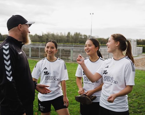 3 piger i sportstøj snakker med en lærer på fodboldbanen