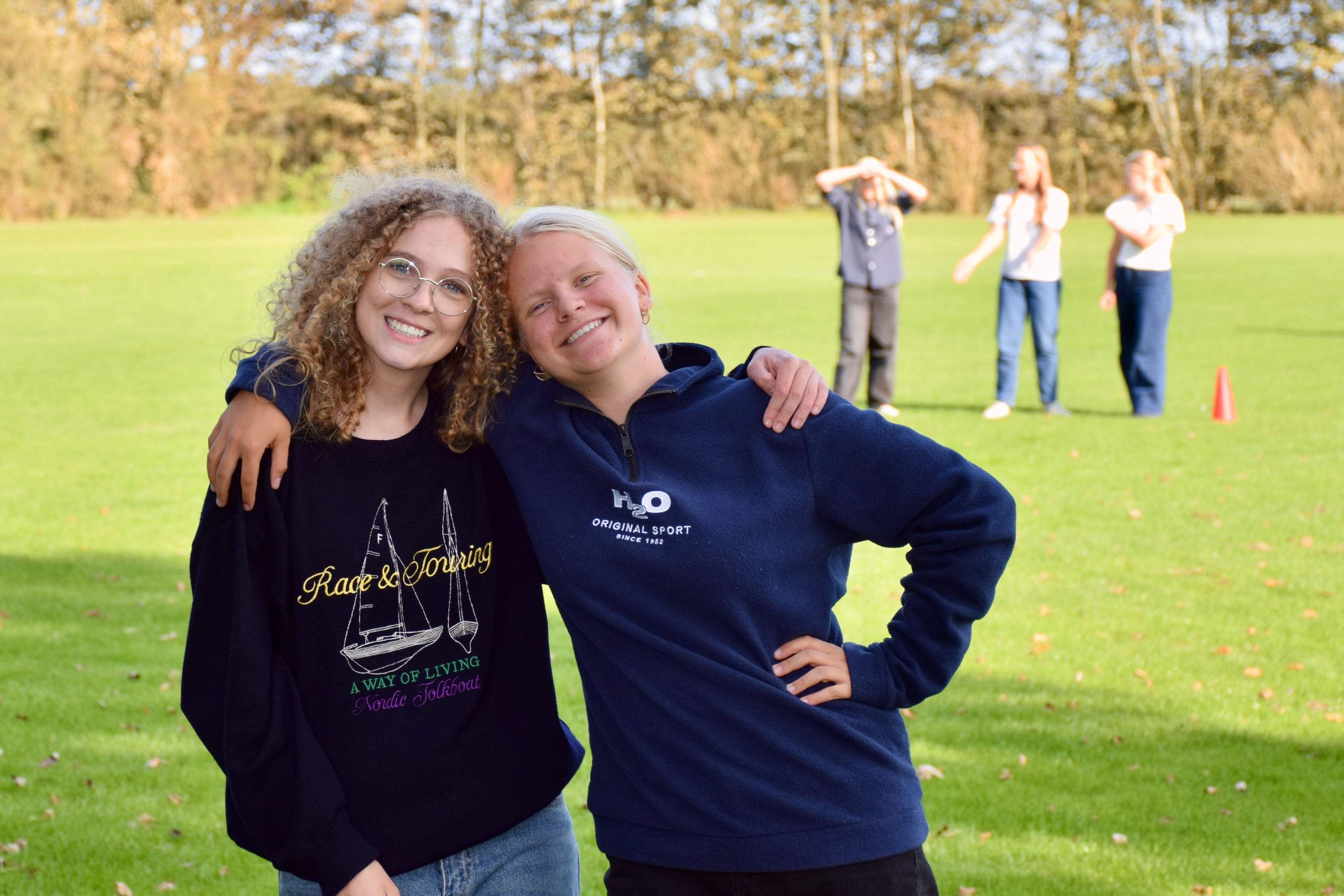 Efterskolernes dag. To efterskolepiger står arm i arm på fodboldbanen, foran en flok som er i gang med at spille rundbold. 