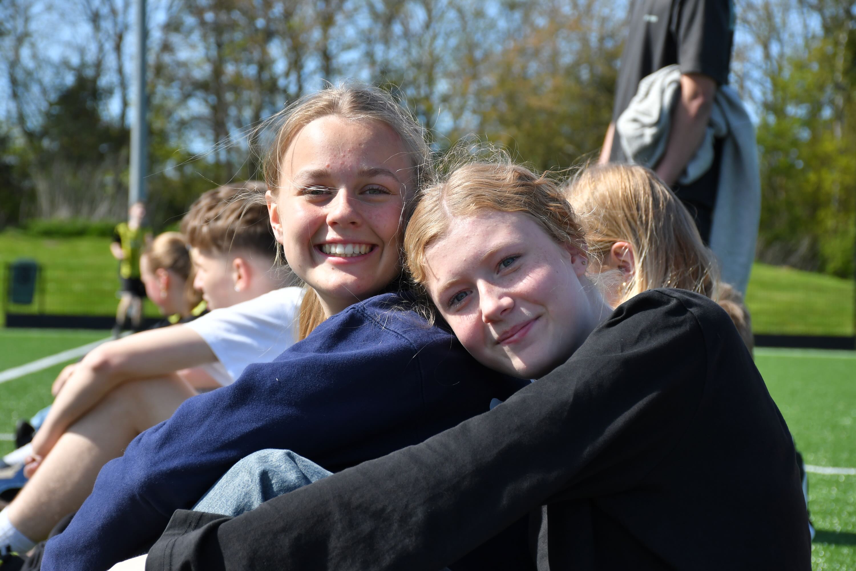 To piger fra Sædding Efterskole sidder og smiler, mens de ser på, at drengene fra efterskolen spiller fodbold. 