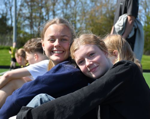 To piger fra Sædding Efterskole sidder og smiler, mens de ser på, at drengene fra efterskolen spiller fodbold.