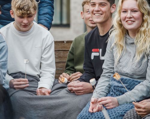 En flok efterskoleelever sidder udenfor ved et bål med tæpper og ristede skumfiduser i hænderne. 