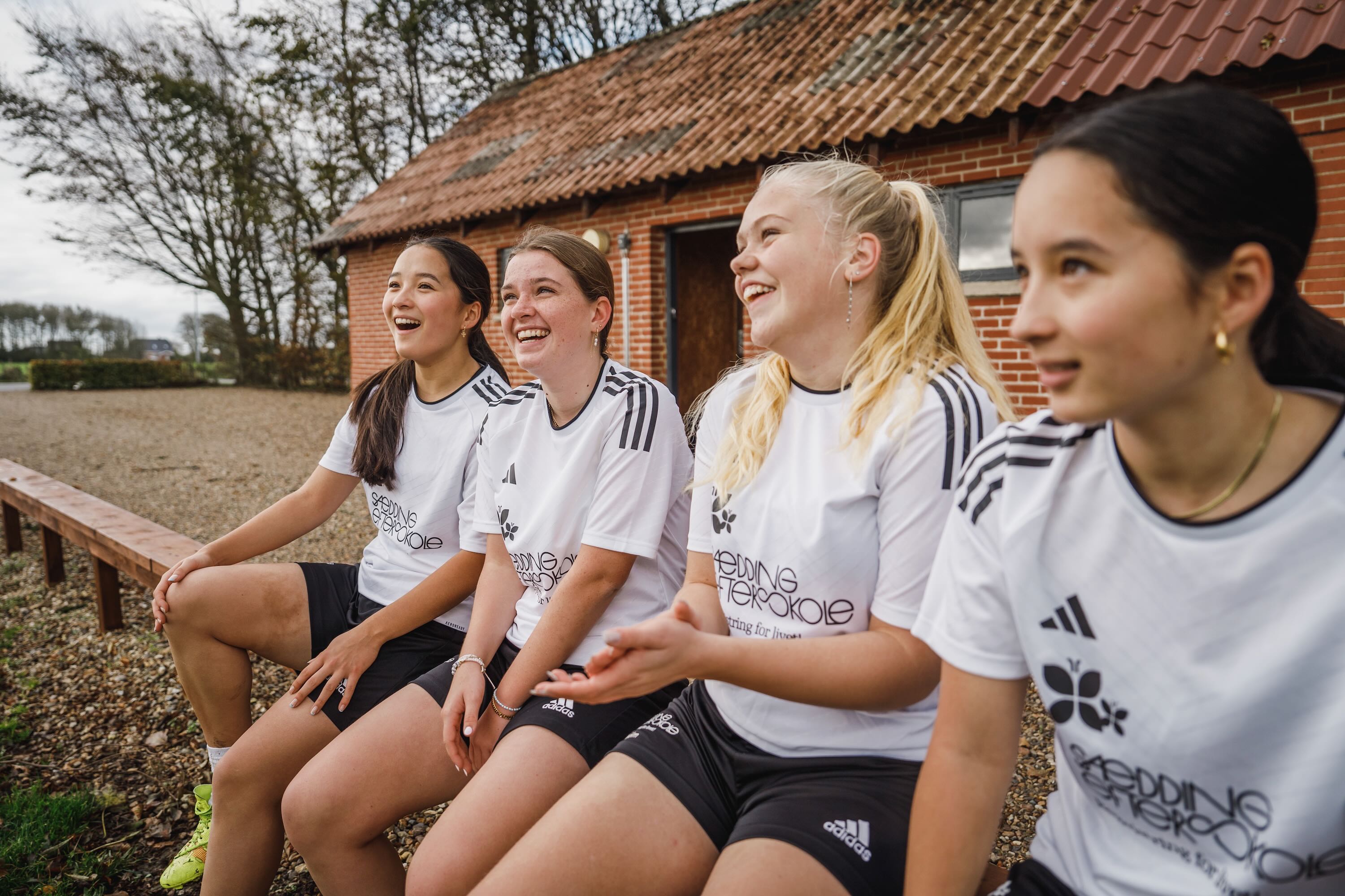 4 efterskolepiger sidder på en bænk på Sædding efterskole i sportstøj og griner. 