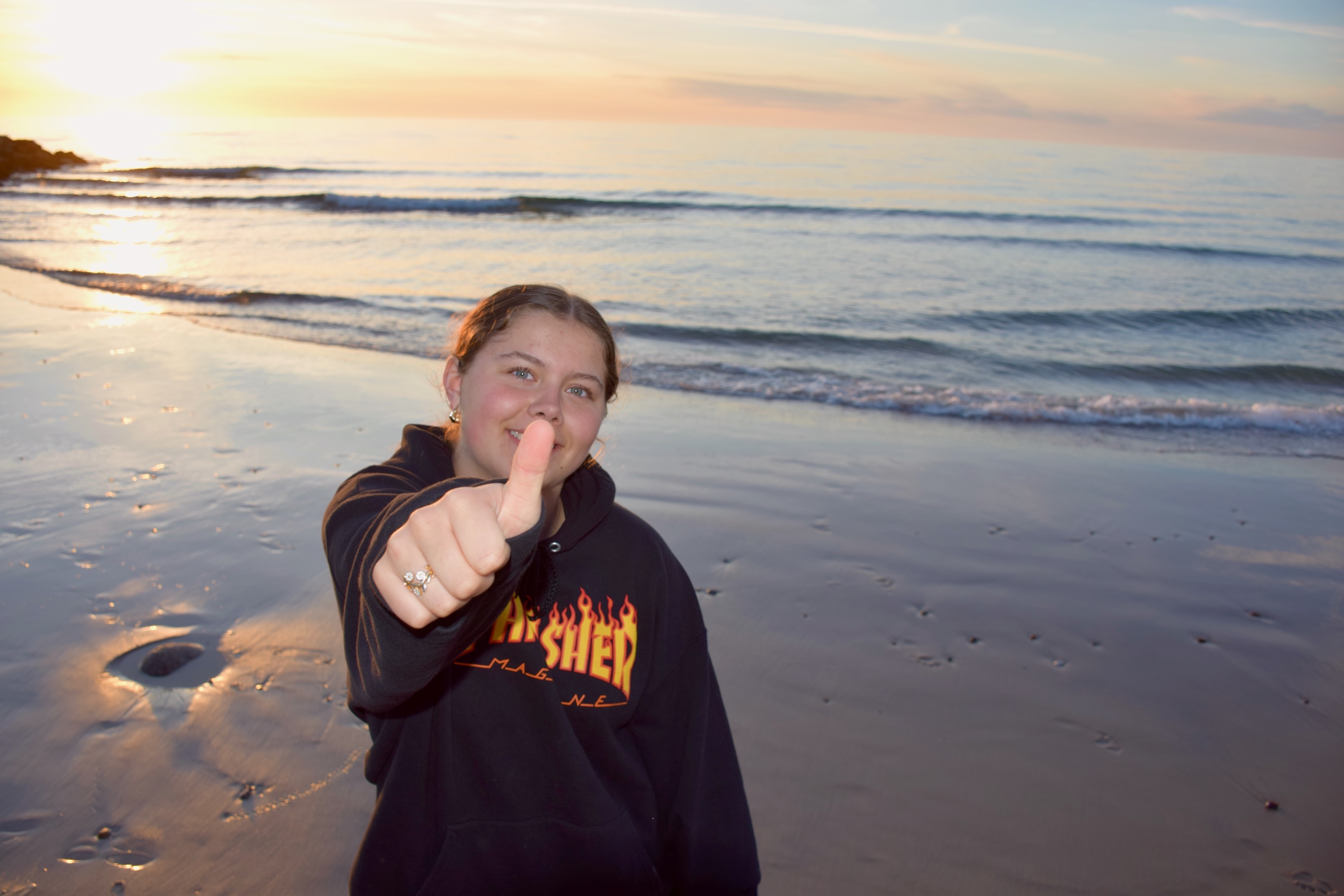 Kristen efterskole i Jylland. En pige fra Sædding Efterskole står på stranden med en solnedgang i baggrunden. Hun smiler og giver thumbs up. 