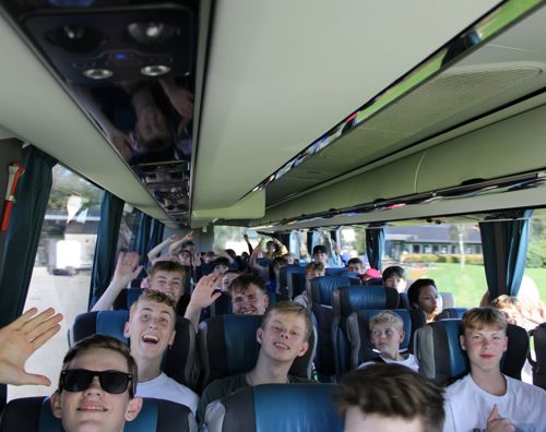 En flok glade efterskoleelever sidder i en bus og smiler og vinker. 