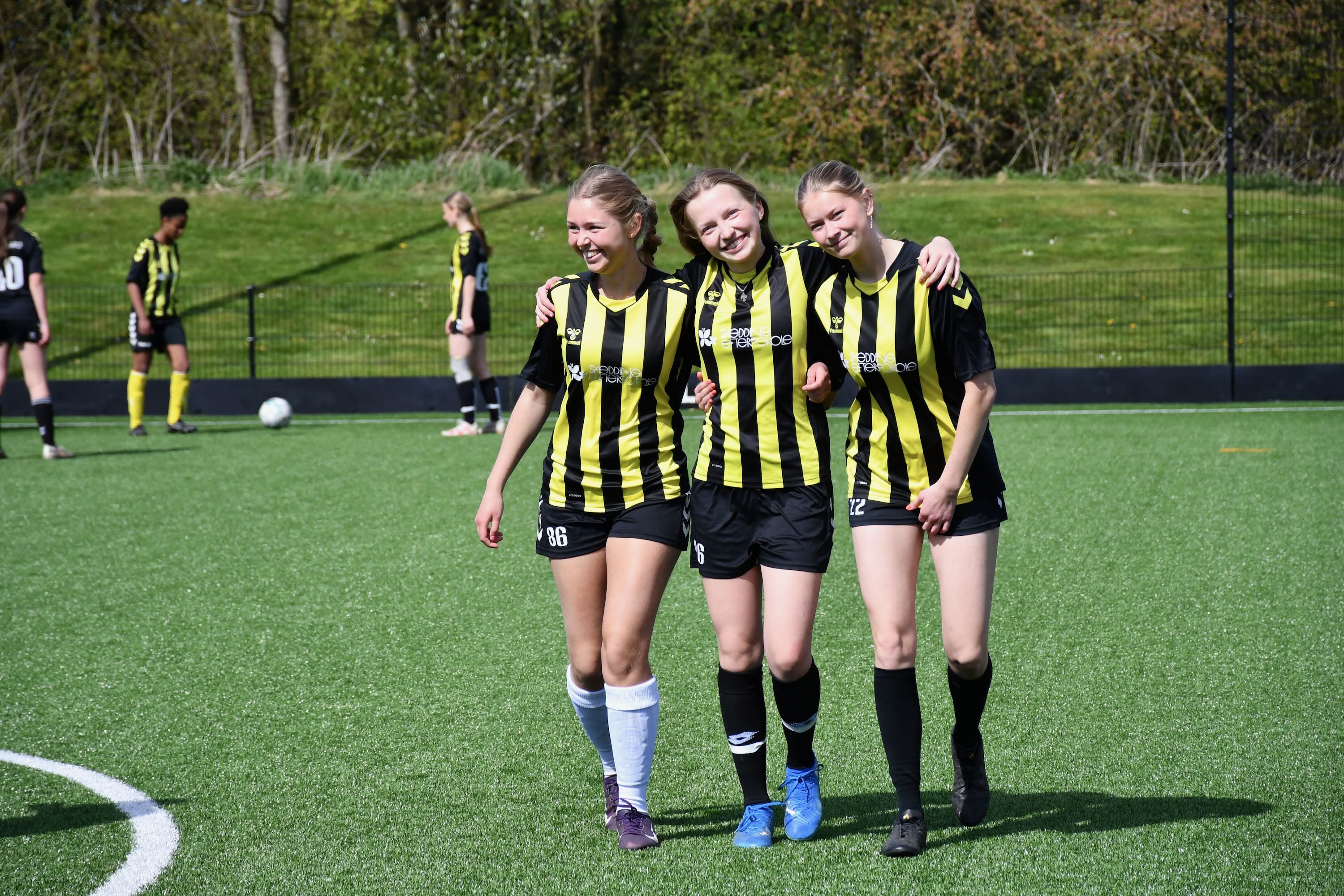 Sport Efterskole | 3 piger går arm i arm i fodboldtøj, smilende hen over fodboldbanen