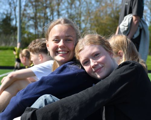 To efterskoleelever fra Sædding Efterskole sidder på en fodboldbane og smiler til kameraet