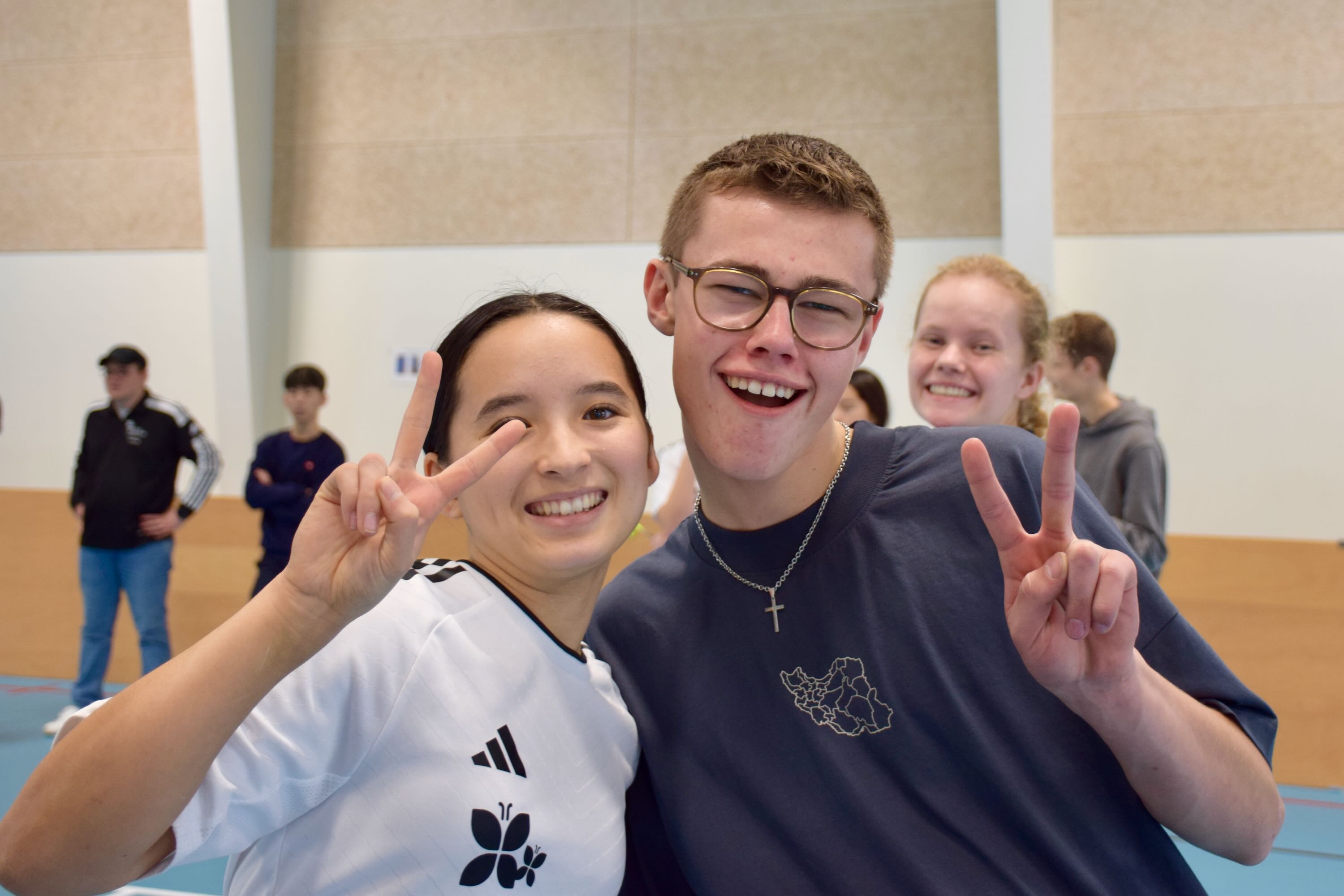 To efterskoleelever står smilende i en hal og giver tegn på sejr med fingrene
