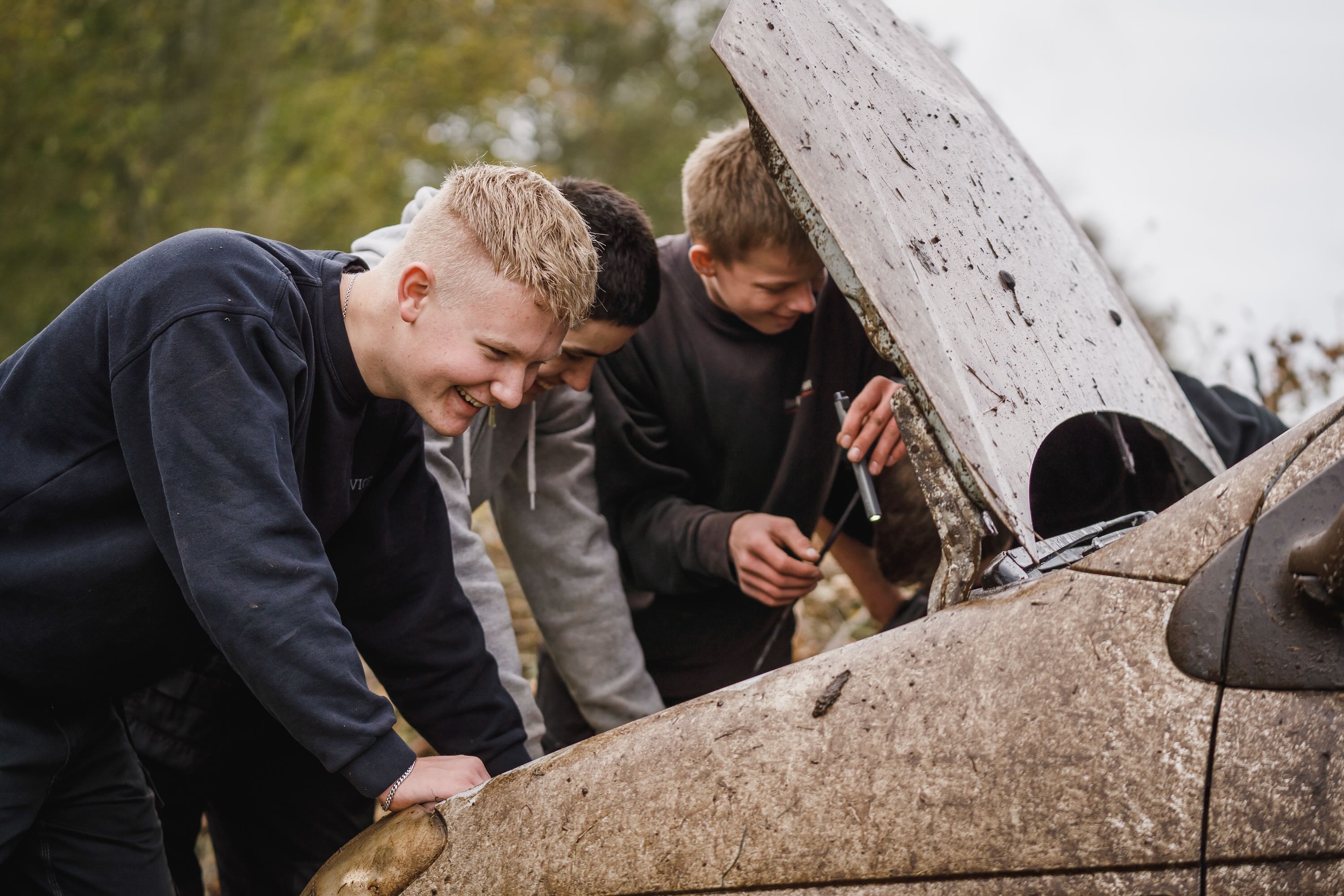 Værksted på Sædding Efterskole | Tre efterskoledrenge står med hovedet nede i en markræsbil og forsøger at få gang i den. 