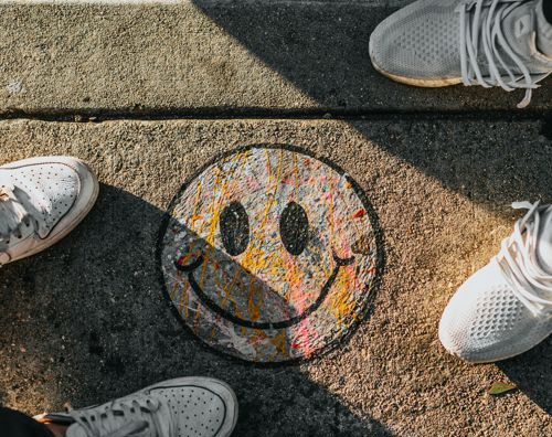 Et smiley er tegnet på asfalten og man kan se tre sæt sko, står rundt om den. 