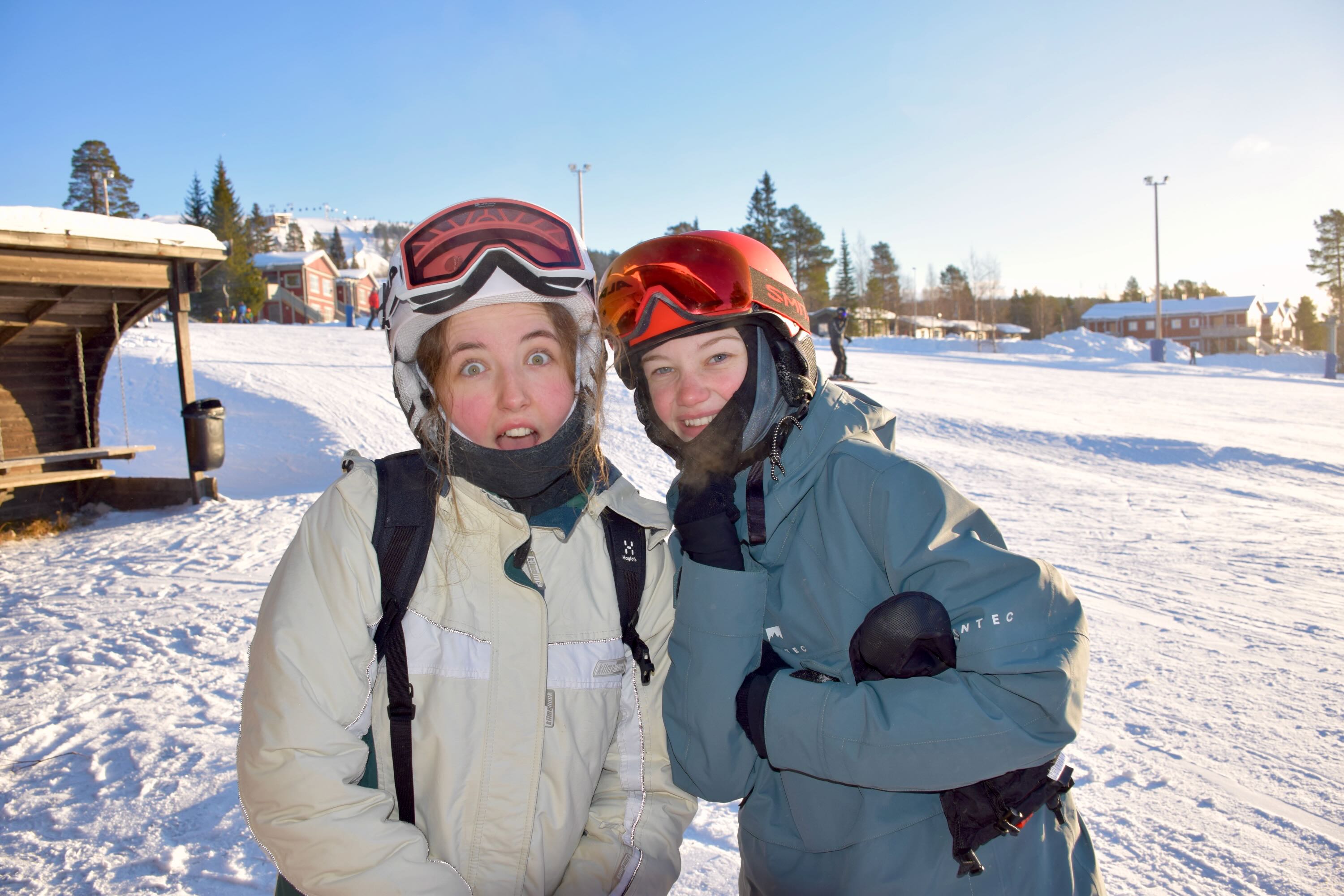 To piger står på en skibakke. Skibrillerne sidder i panden og de smiler. 